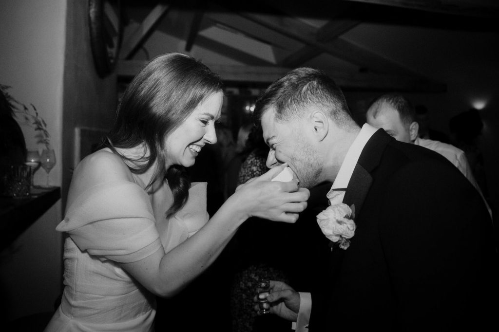A bride holds out a wedge of lime in both hands as her new husband bites down on it and winces with the sourness; he holds an empty shot glass in his hand. His wife smiles in pure delight.